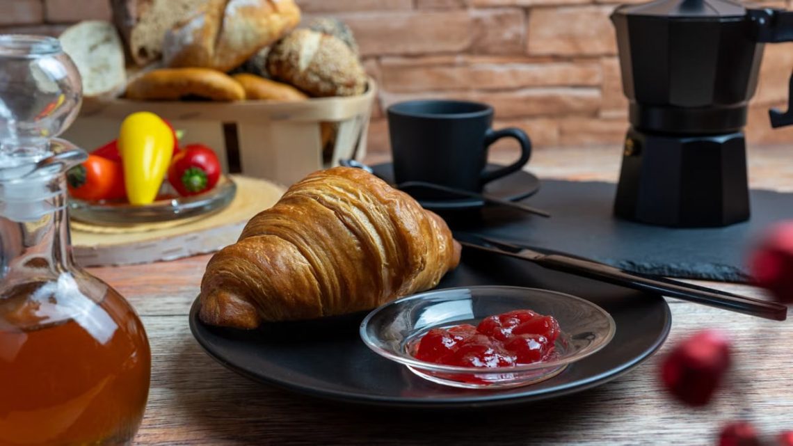 A cozy breakfast setting featuring a croissant with jam, coffee, and a variety of bread.