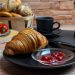A cozy breakfast setting featuring a croissant with jam, coffee, and a variety of bread.